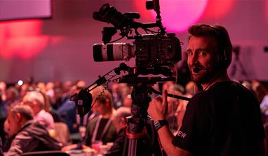 A man operating a big camera and wearing a headset smiling at the camera with a thumbs up.