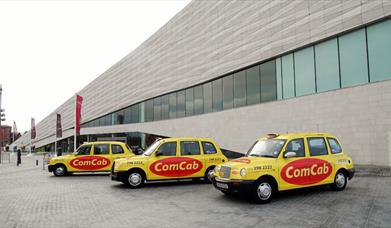 Three taxis with 'ComCab' logo on the side. The taxis are yellow with a red logo and are situated outside Museum of Liverpool.