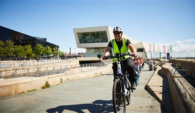 A man in a high vis jacket and helmet riding a bike along Liverpool Pier Head