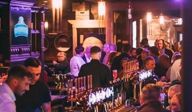 A traditional pub is bathed in purple lighting. There are a number of beer taps on the bar. The walls are traditionally cladded.