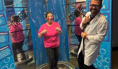 A lady in a pink t shirt stood inside a bubble. A man in a white lab coat smiling