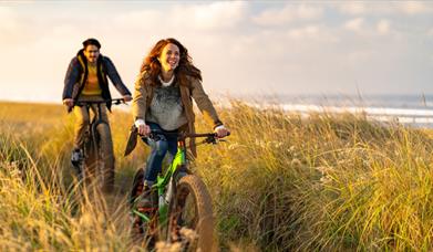 Man and woman cycle through grassland along the coast.