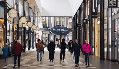 People walking through a selection of stores with large windows and black and white tiled floor. At the end of the corridor of stores you can see a la