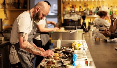Two male chefs preparing food on an open kitchen next to a bar with people watching on