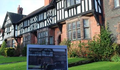 A black and white and red brick Victorian building in Port Sunlight. The building is well kept and has lush green grass and tall wall flowers.