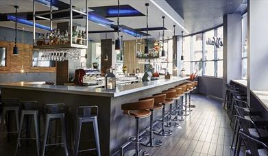 A modern bar with located in the centre of the restaurant. There are metal bar stools lined up against the modern bar which has hanging wine glasses a