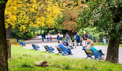 Families sit on benches within Sefton Park during Autumn time. The trees have yellow, red and green leaves.