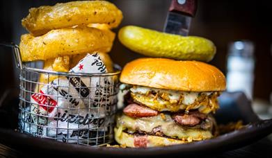 A large burger with a gherkin on the top next to a small basket of onion rings.