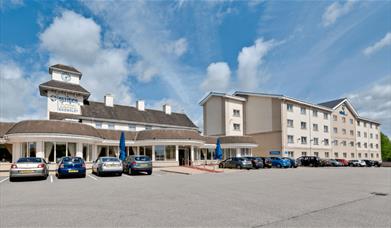 An exterior image of The Suites Hotel and Spa with cars parked outside and a bright blue sky.