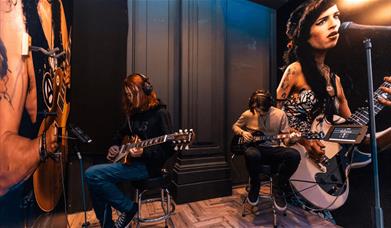Two people sitting playing guitars with a picture of Amy Winehouse in the background.