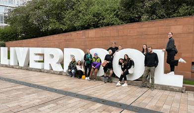 A group of people sitting and standing on a large white LIVERPOOL sign.