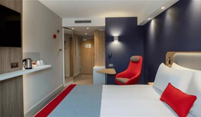 A double bed with a red pillow and a red chair with a large tv on the wall.