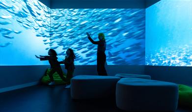 An adult and two children inside a room with fish swimming projected onto the walls.