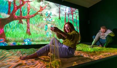 A woman sits playing in a large sand pit in a darkened room with a colourful, projected nature scene on a screen behind
