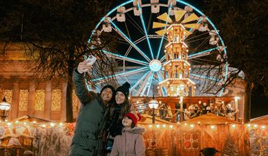 People taking a selfie in front of the Christmas market in front of the Neo-classical building that is covered in fairy lights